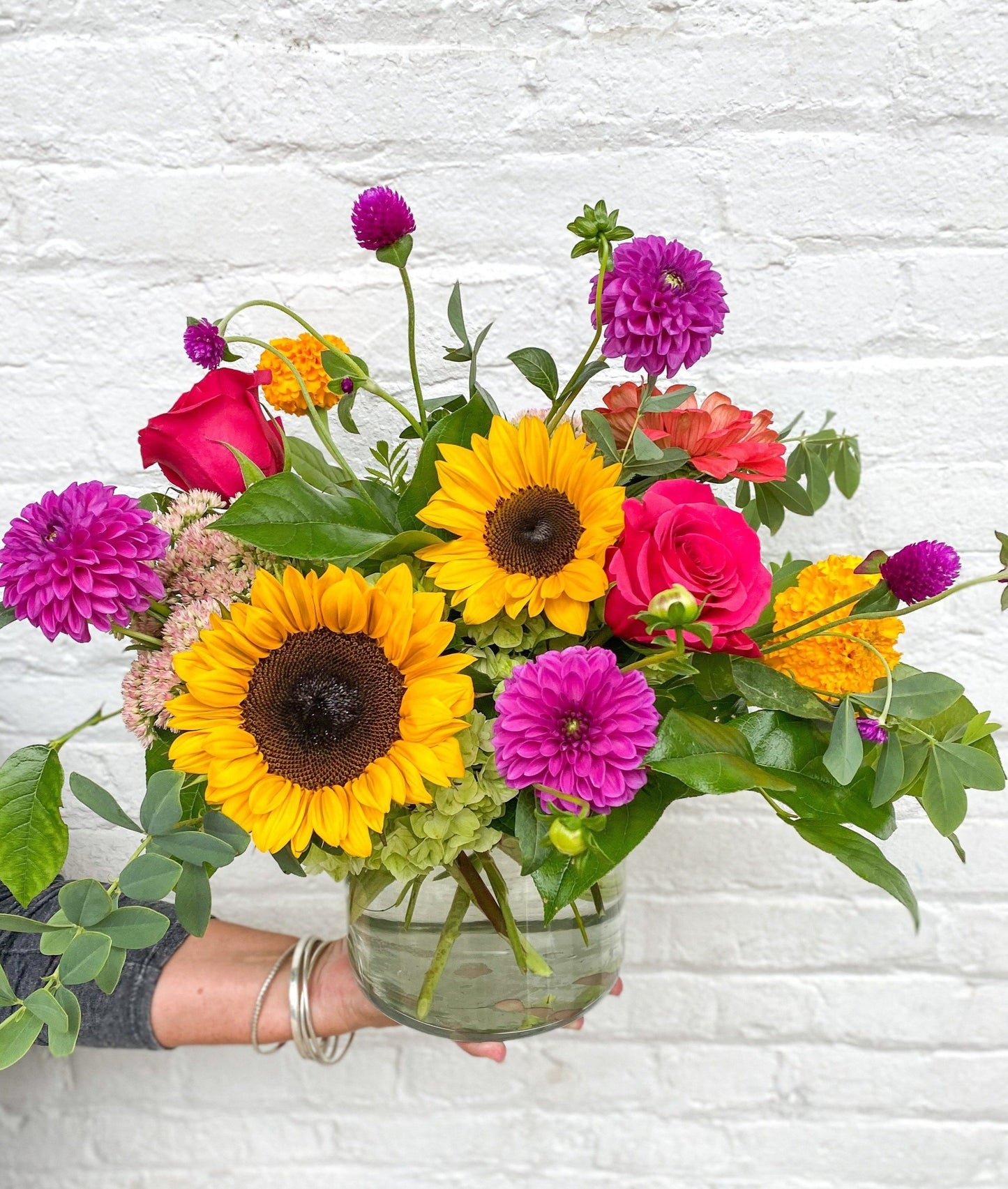 Bouquet of colorful flowers including sunflowers and roses held against a white brick wall.
