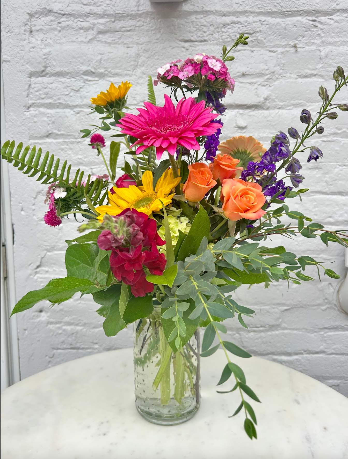 Bouquet of colorful flowers in a clear vase against a white brick wall.
