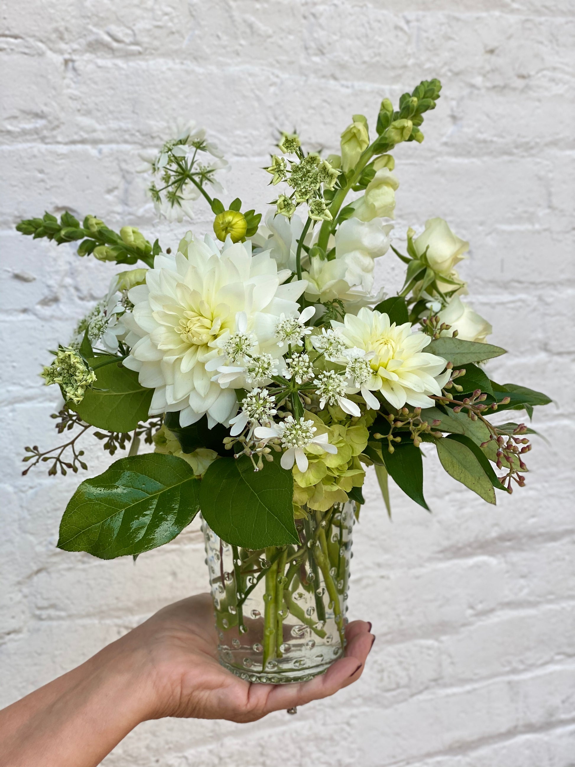 Bouquet of white and green flowers in a clear vase held against a textured white background